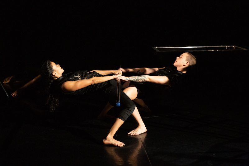 Two dancers hold hands facing one another and hinge their upper bodies parallel to the floor.